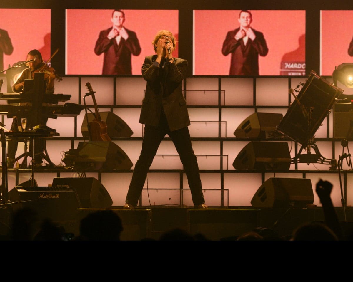 A singer performs with a microphone, legs apart, in front of screens showing his image
