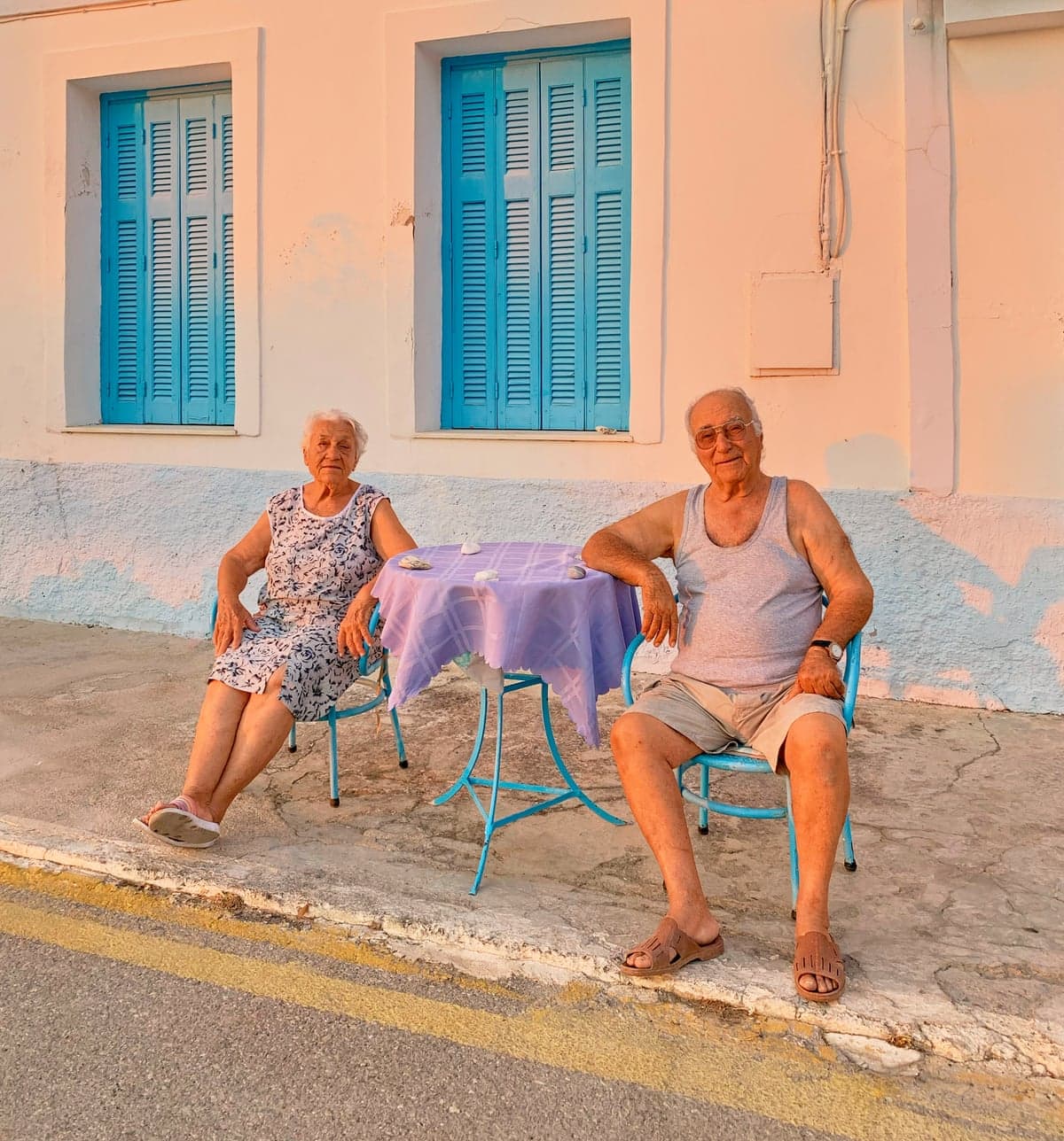 An elderly Greek couple sitting on light-blue chairs turned towards the camera on a pavement in front of a house with light-blue shutters. They are sitting either side of a light-blue table covered by a light-purple tablecloth