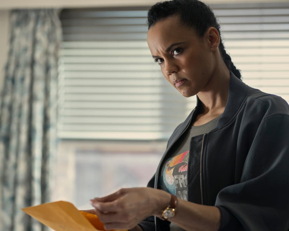 A young woman wearing a T-shirt and dark bomber jacket looks concerned while holding a yellow envelope