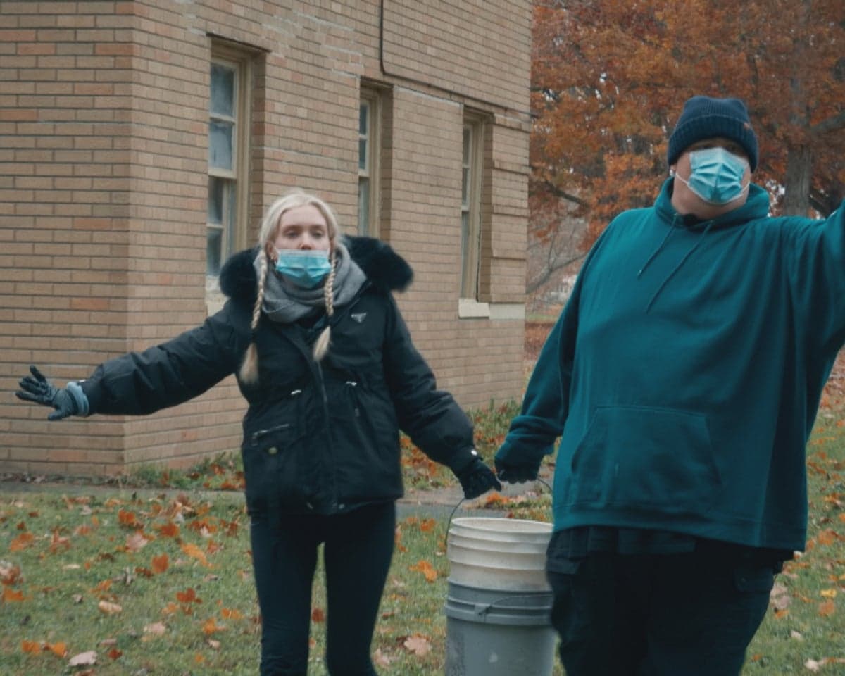 A woman in fur-trimmed hooded dark jacket and plaited blond hair and a man in a large turquoise hooded sweatshirt, both wearing blue facemasks, hold the handle of a stack of buckets with their other arms stretched out to the side - in front of a modern bricked building with three windows with autumn leaved trees to left and leaves on the grass they're standing on