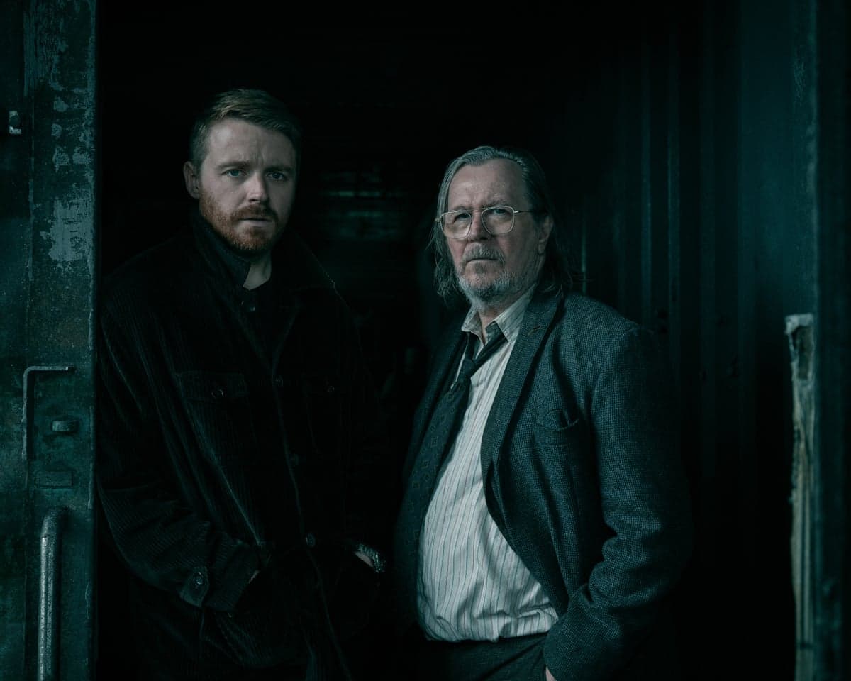 Jack Lowden and Gary Oldman stand together in a dim, shadowy doorway in Slow Horses.