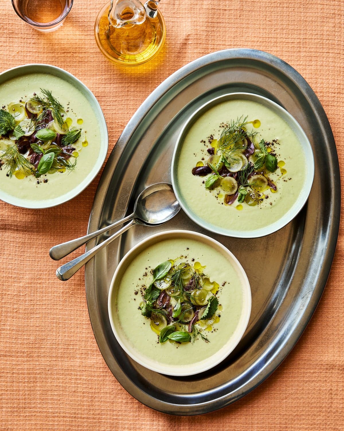 Three bowls of pale green soup topped with olives, herbs, and oil on a serving tray. AKA Alexina Anatole's cucumber and avocado gazpacho.