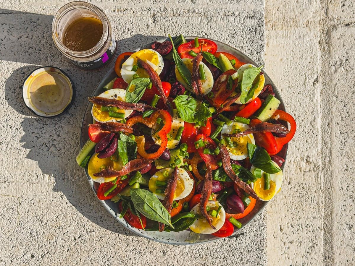 Overhead view of a colourful salad with eggs, anchovies, tomatoes and olives, beside olive brine dressing jars.