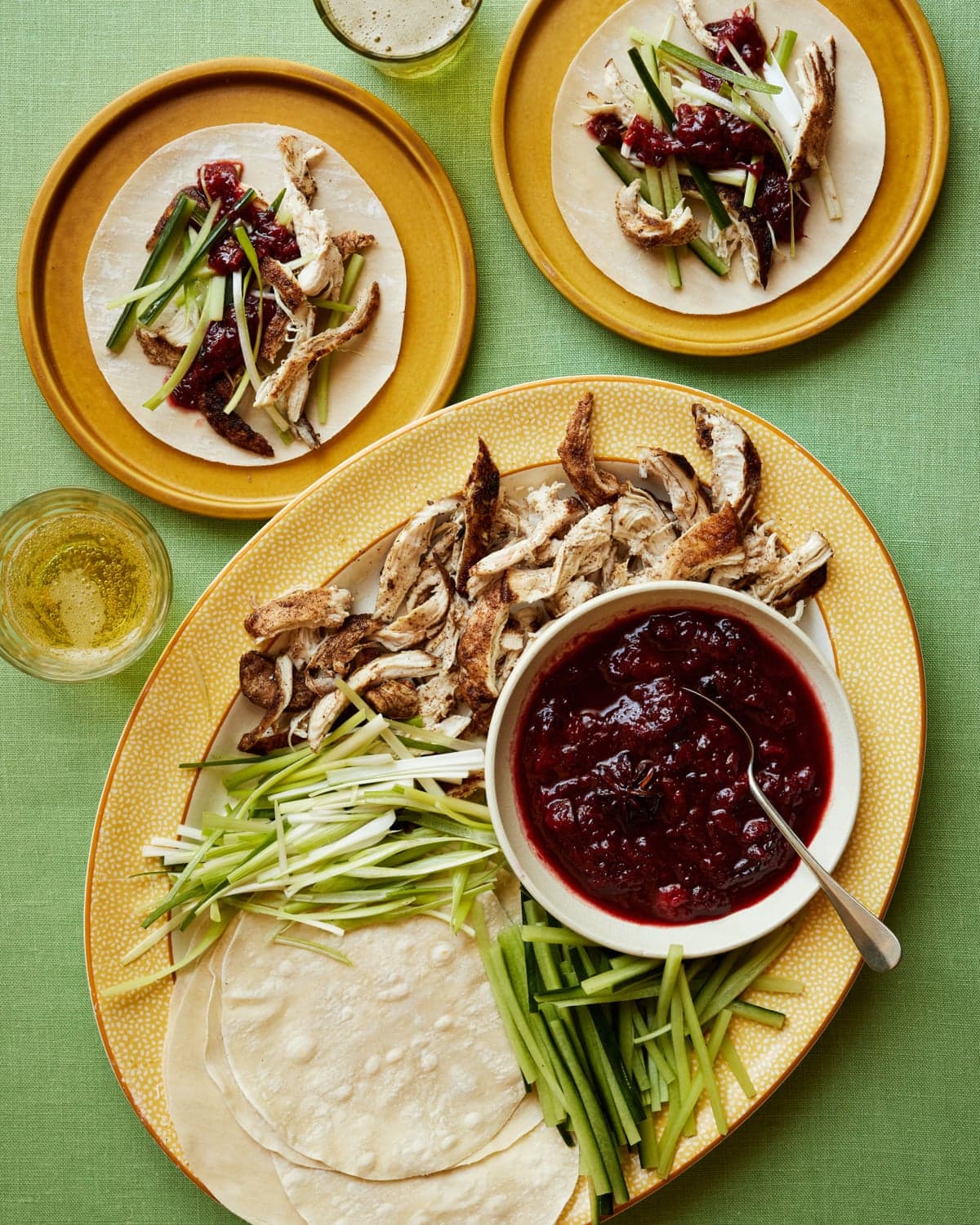A platter displays pancakes, shredded chicken, cucumber strips, and a bowl of dark plum sauce alongside assembled wraps