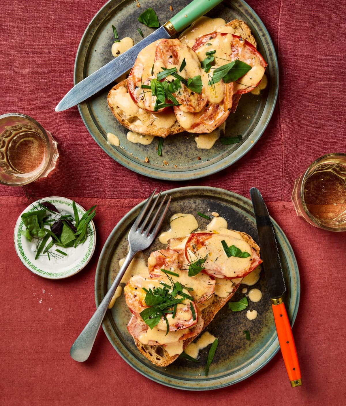 Two plates of toast topped with sliced tomatoes, creamy sauce, and fresh herbs, with cutlery and drinks nearby