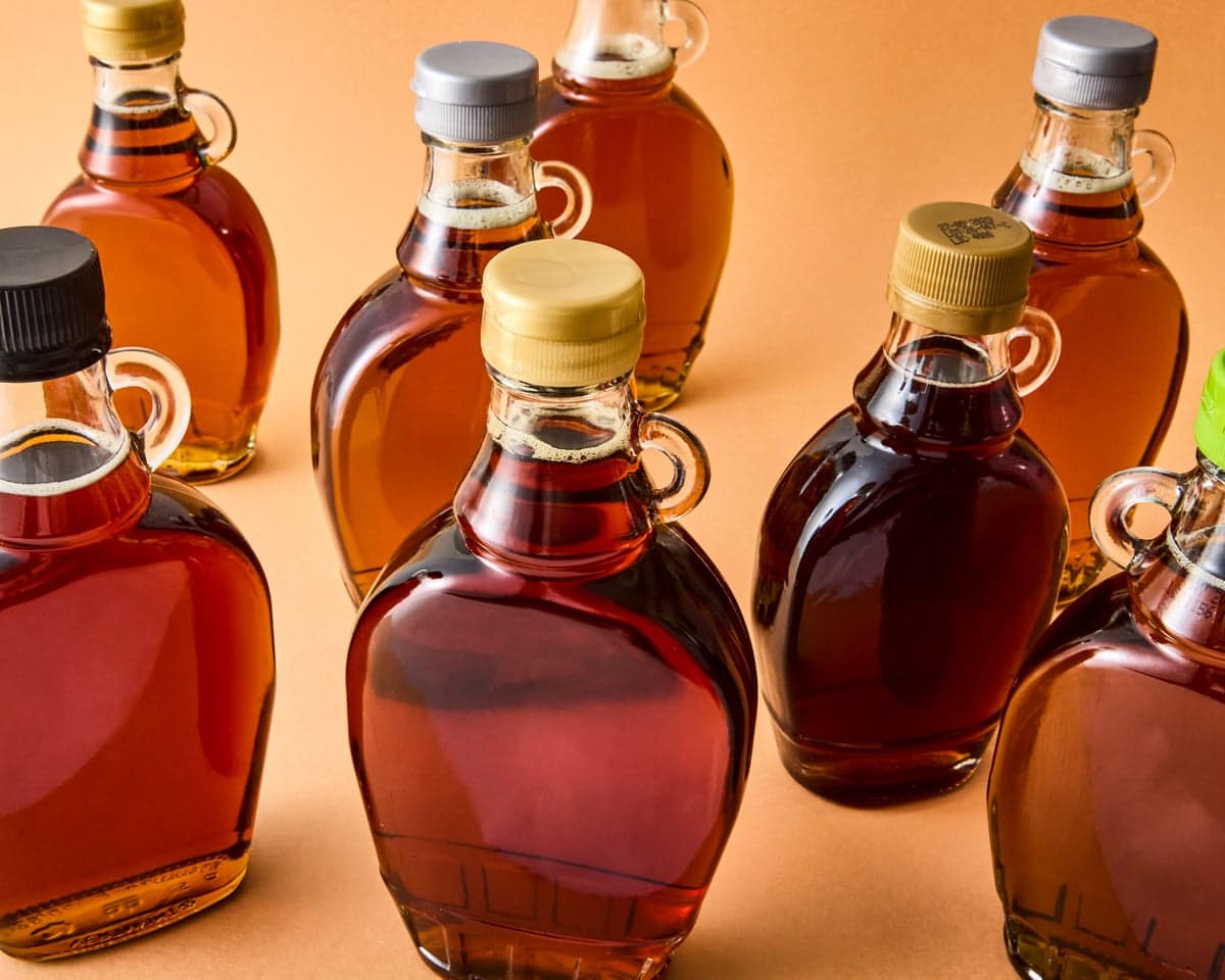 Seven glass jugs filled with amber maple syrup in varying shades stand on an orange surface