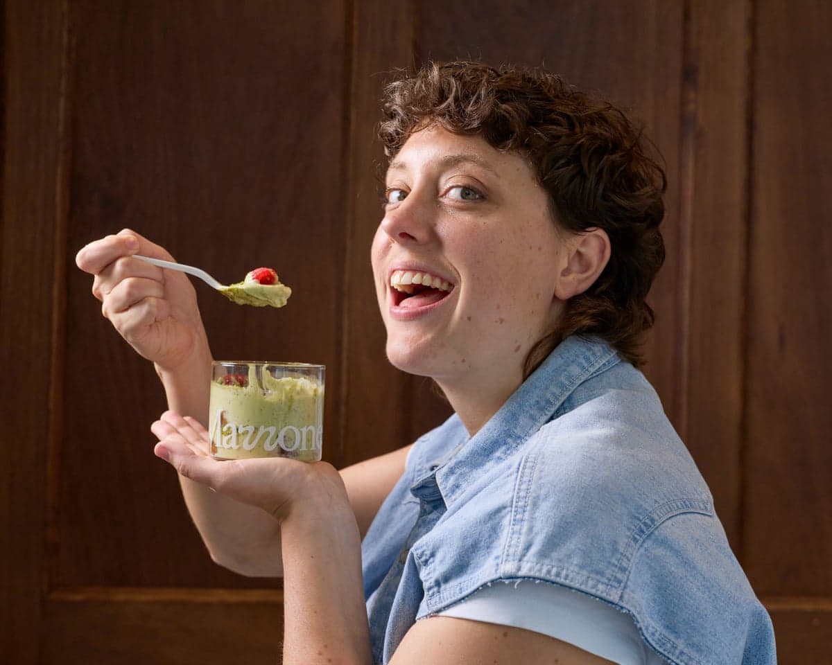 A head and shoulders of a young woman with short curly brown hair wearing a denim short-sleeved shirt - side on to camera looking round smiling with mouth wide open with a pot of green cream in a glass pot with the words 'Marrone" in left hand and a spoon with green cream and a raspberry on top in her right - poised to eat