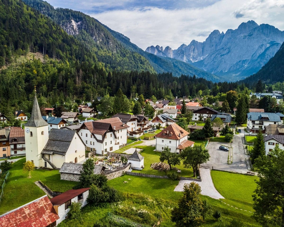 View of a village with white houses and a steepled church, flanked by forest and mountains