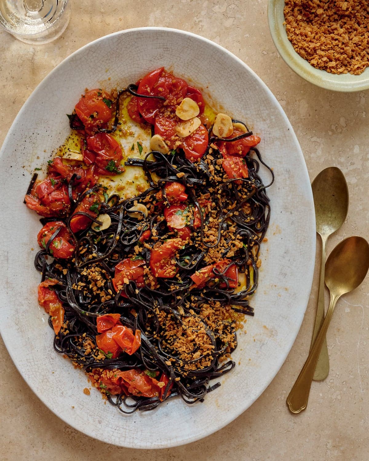 Chris Leach's squid Ink spaghetti with anchovies, cherry tomato and pangrattato on a platter with cutlery and drinks nearby