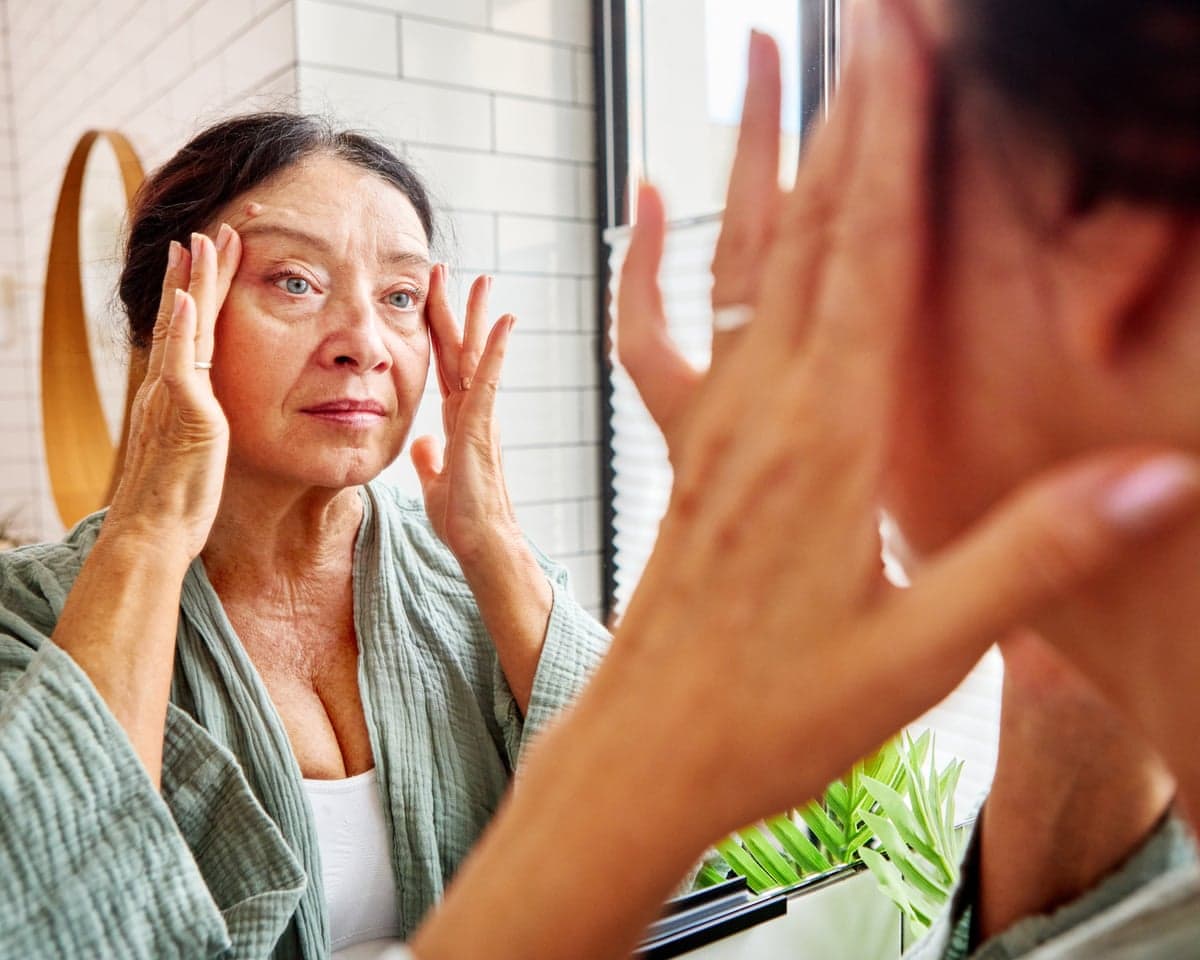 A senior woman touches her fingertips to her temples while looking at her reflection