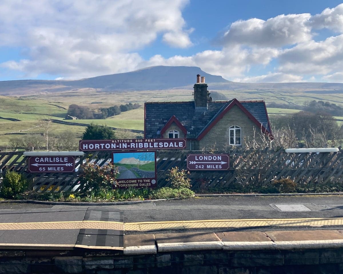 A railway platform backed by Yorkshire hills, with station sign and signs with arrows pointing to Carlisle and London in the foreground