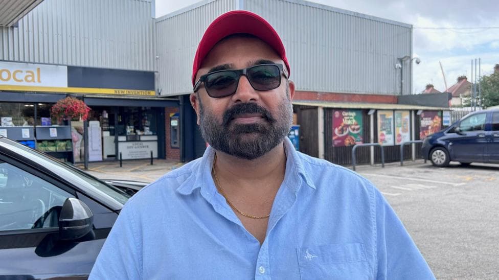 Arvander Patel stands on the petrol station forecourt. Behind him is a grey building and a number of cars. He is wearing a red baseball cap, black square-framed sunglasses, a gold chain and a light blue shirt.