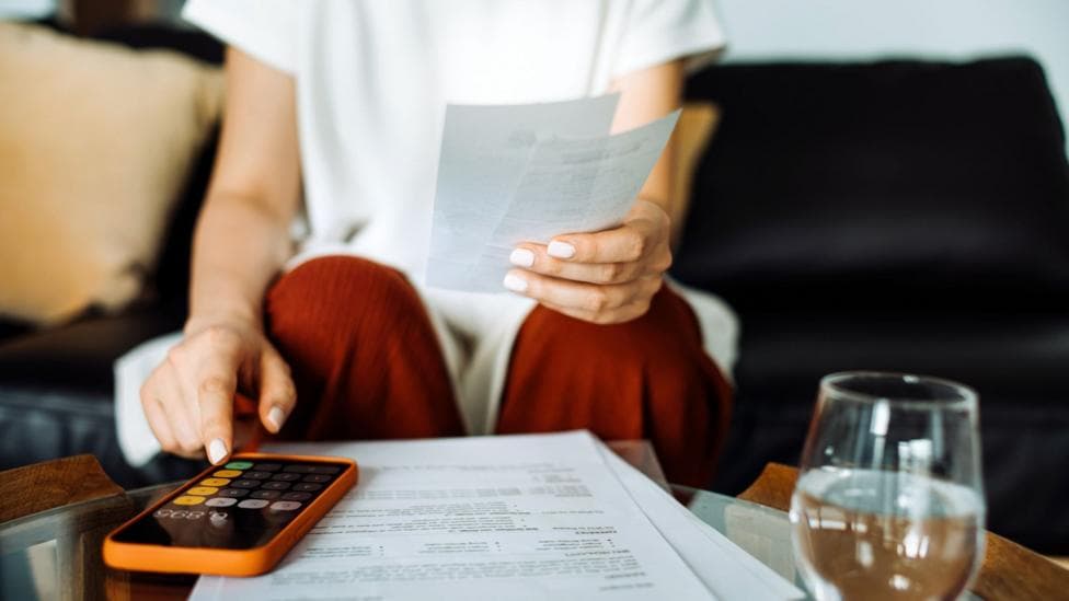 A woman sits down with a glass of water and her mobile phone. She is making calculations looking at several pieces of paper in front of her.