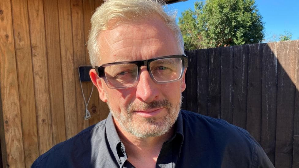 A man with white/blonde short hair, grey framed glasses and stubbly facial hair. He is wearing a black polo shirt with two buttons undone. He is stood in front of a brown fence to his right and a light brown shed to his left.