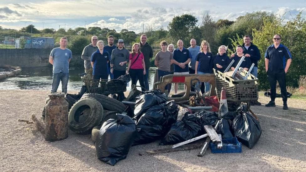 A team of 16 divers, dressed in everyday clothes such as jeans and t-shirts standing in front of a large pile of fly-tipped waste. The pile includes a number of old car tyres, a shopping trolley, a large milk churn, many large full black bags, a Highways Agency barrier, planks of wood and what looks like a side table upside down.
The team is smiling proudly with the River Nene just behind them, and the sky is blue with some white wispy clouds.