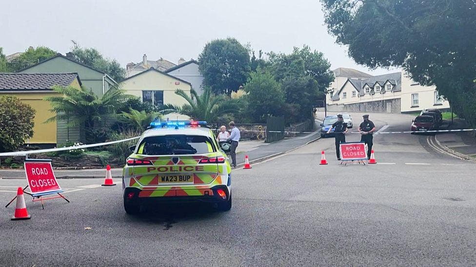 A police officer stands by his police car on the side of a road, which is cordoned off with police tape and orange cones.
