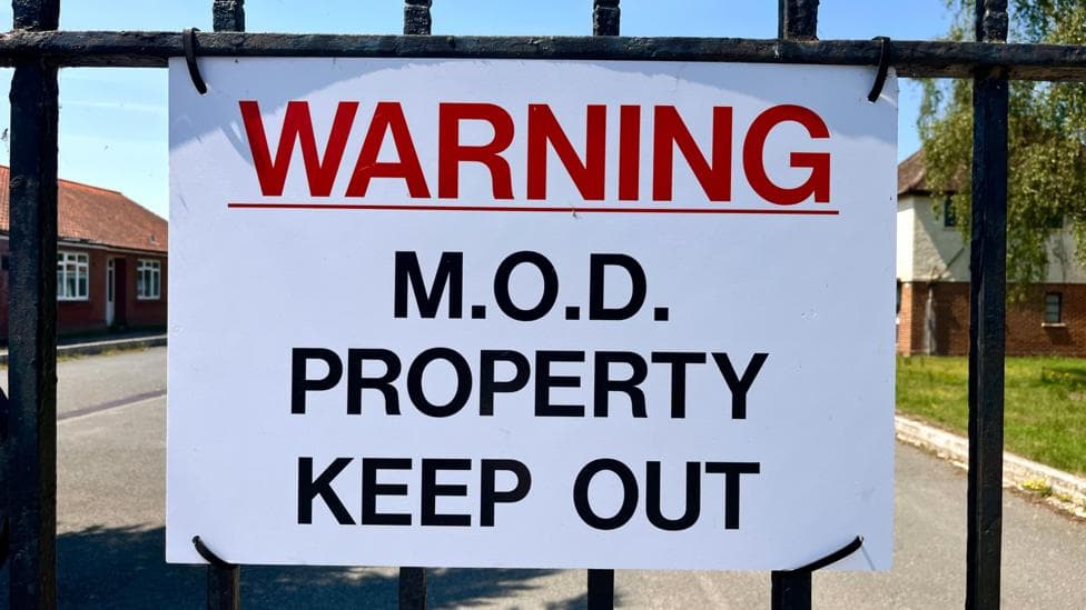 A sign says: "WARNING: MOD PROPERTY, KEEP OUT". It is white with red and black writing and is on a metal gate. Beyond it is a road and some buildings.