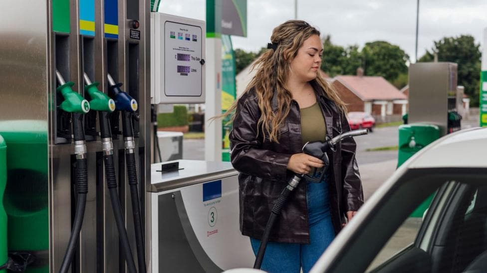A stock image showing a woman wearing blue jeans, a green top and a black leather jacket. She has long and wavy brown hair and is filling up her car at a petrol station.