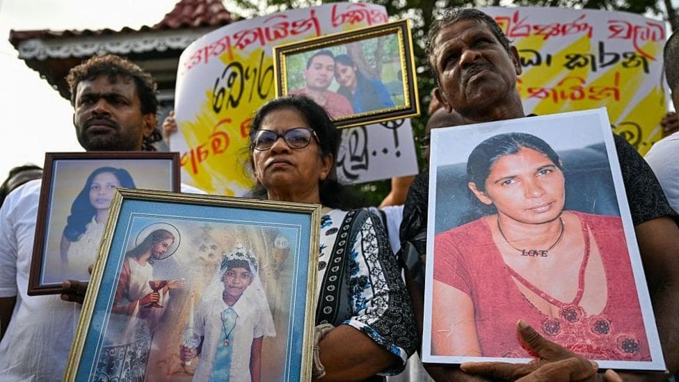 People stand at a public protest holding large framed photographs of several individuals. One person in the foreground holds a portrait of a woman, while others display photos of different people, including a framed childhood image and a picture of a couple.