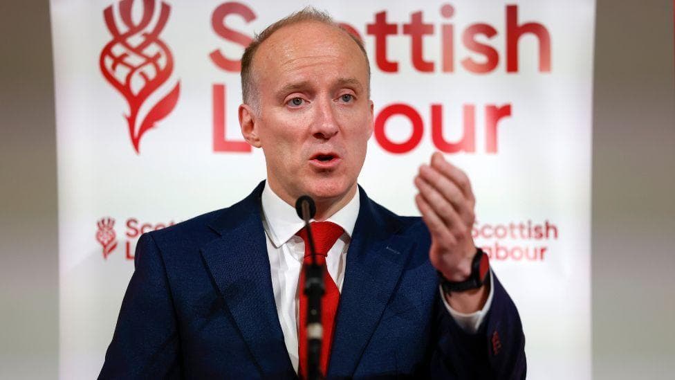 Michael Marra, who has thinning ginger hair, speaks into a microphone in front of Scottish Labour branding. He is wearing a business suit
