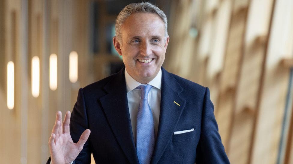 A man with short grey hair waves and smiles. He is wearing a business suit