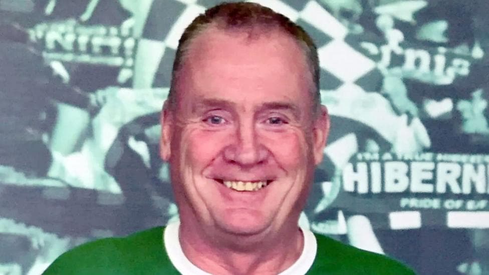 A man is smiling at the camera. He is standing in front of a poster that says Hibernian football club. He is wearing a green and white t-shirt. He has short brown receding hair.