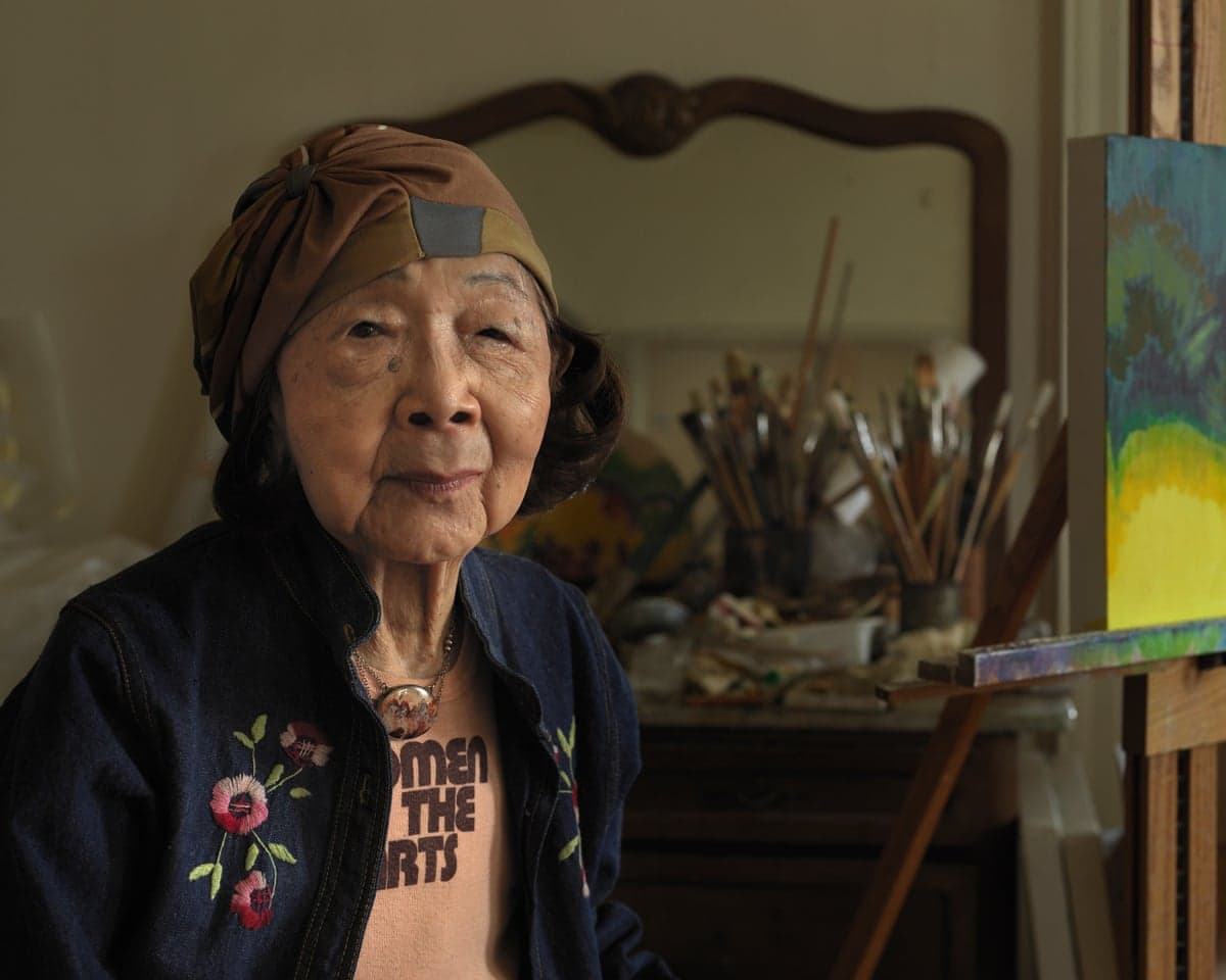 Artist Ce Roser, in a headscarf and denim jacket, smiles near her studio's painting supplies
