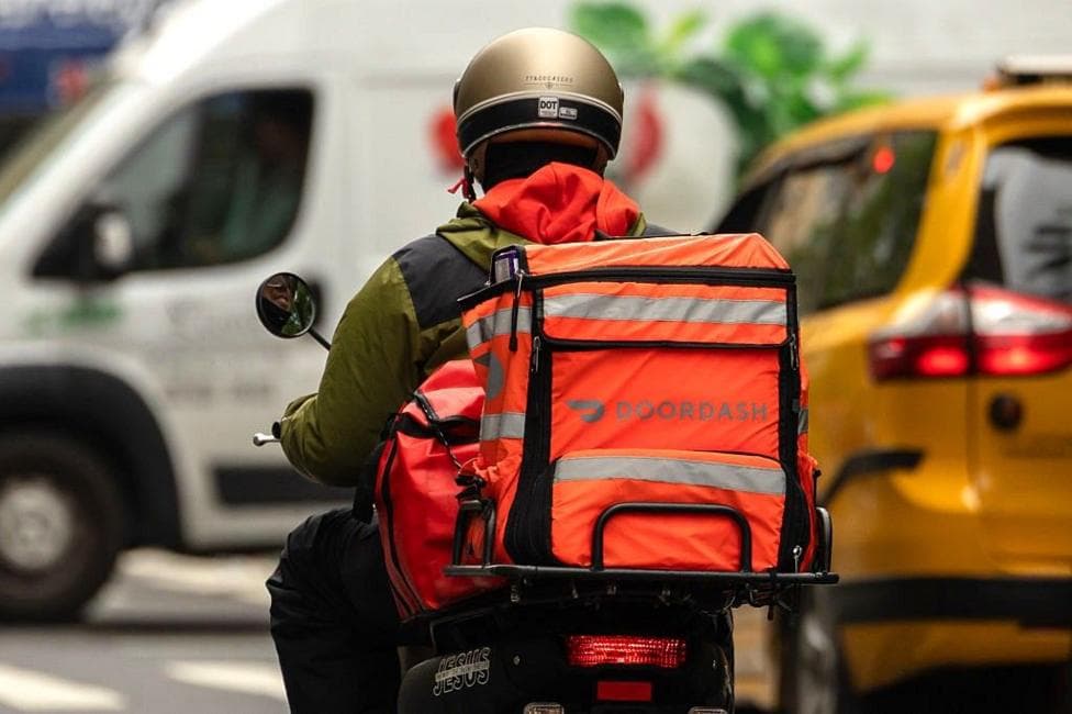 Person rides a moped with an brightly-coloured bag attached to the back with the logo reading Doordash