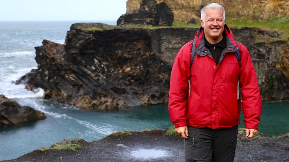 Derek's wearing a red waterproof jacket as he stands on a coastal hilltop overlooking the sea below