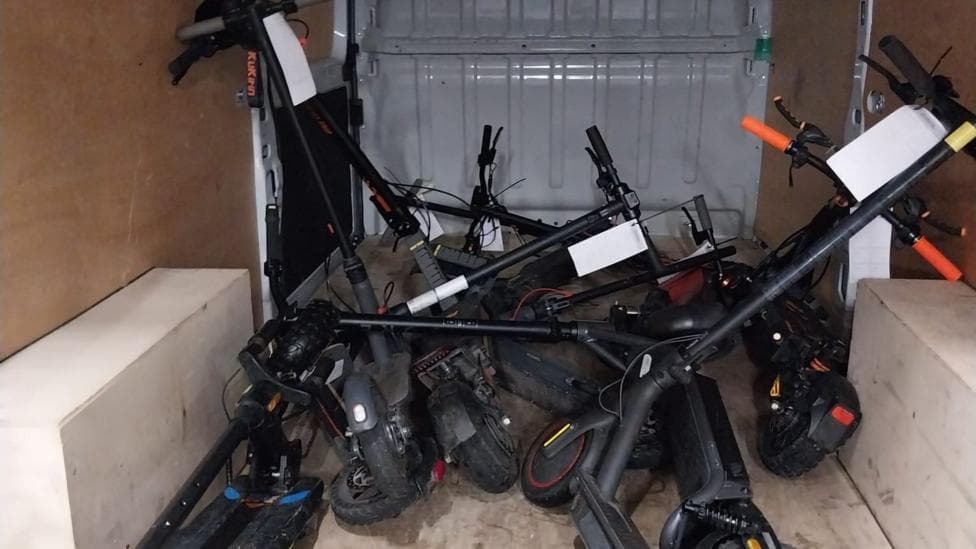 Black e-scooters in the back of a van. They are piled on top of each other with white tags on them.