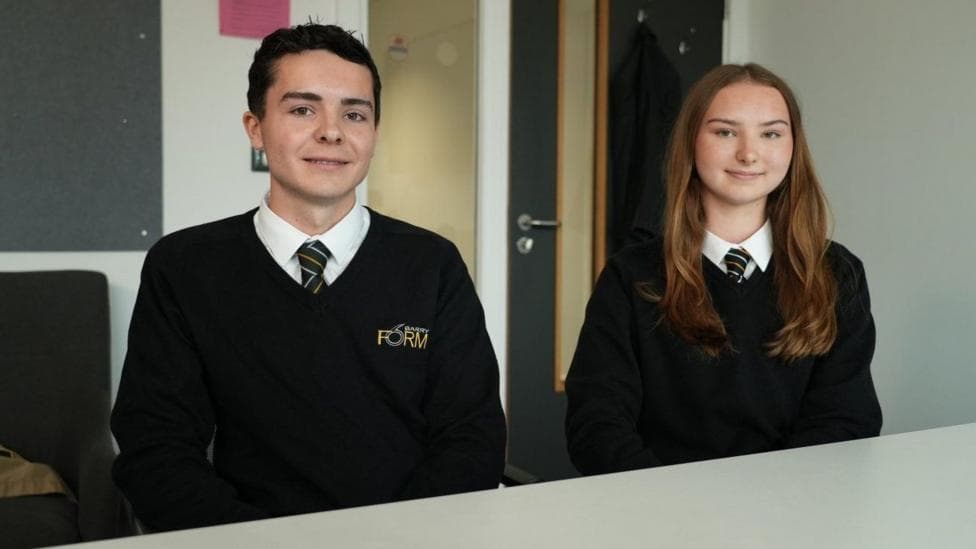 Two students are sat at a desk, looking at the camera. Jay, a 17 year old boy, is on the left, Amelie, a 17 year old girl, is on the right. They are both wearing black school uniform jumpers for Barry Sixth Form.