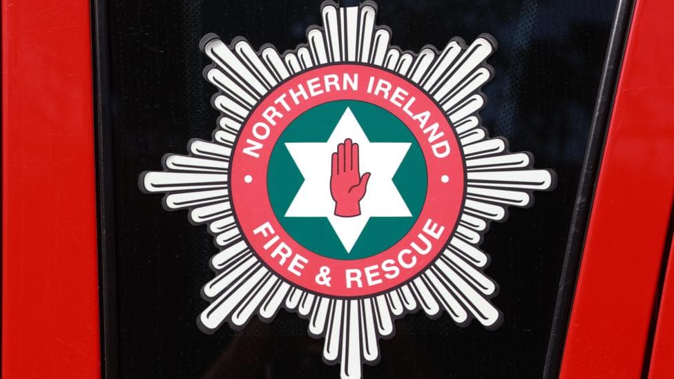 A crest of the Northern Ireland Fire and Rescue Service is shown on the side of a fire engine. The crest has a red hand in the centre surrounded by a white star and a green circle extending around it. The name of the organisation is encased further out around the crest.