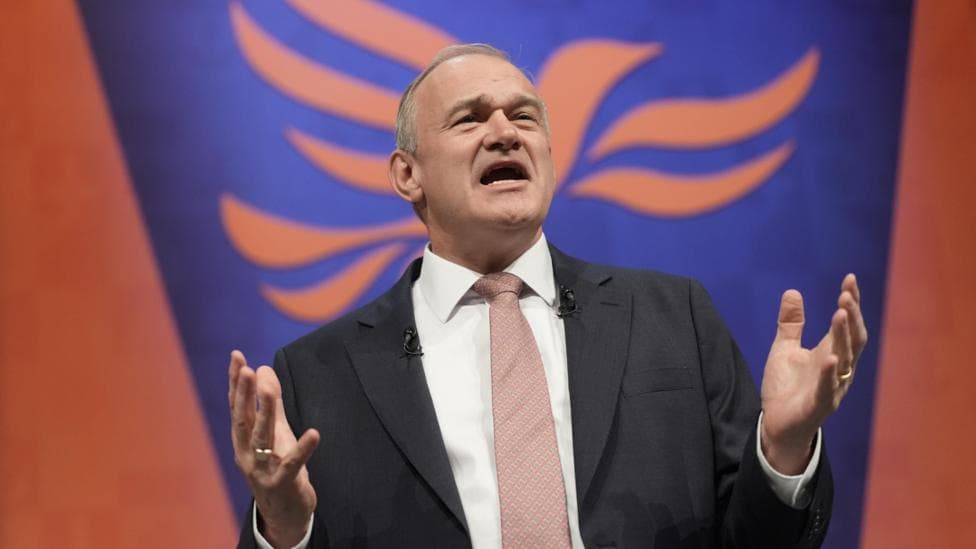 Sir Ed Davey at the Liberal Democrat party conference. He is pictured against a blue and red background, and he is gesturing with his hands as he makes his speech.
