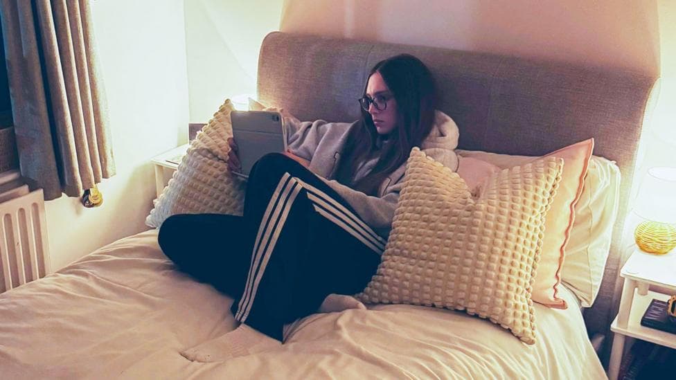 Anna-May, who has long brown hair and wears glasses, is propped up on lots of cushions on her bed, which has a light grey headboard. She is watching an iPad which she is holding in her hands, wearing tracksuit bottoms and a hoodie. There are bedside lights on tables on either side of her bed.