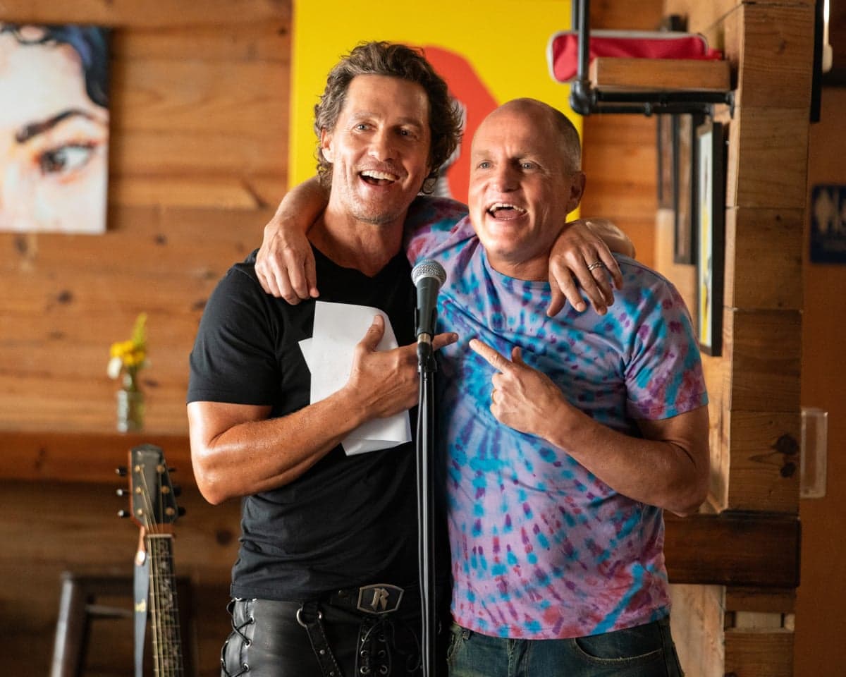 Two middle-aged men with arms round each other and appearing to be singing or shouting happily one in black T-shirt and one in multi-coloured T-shirt in a pine-panneled room