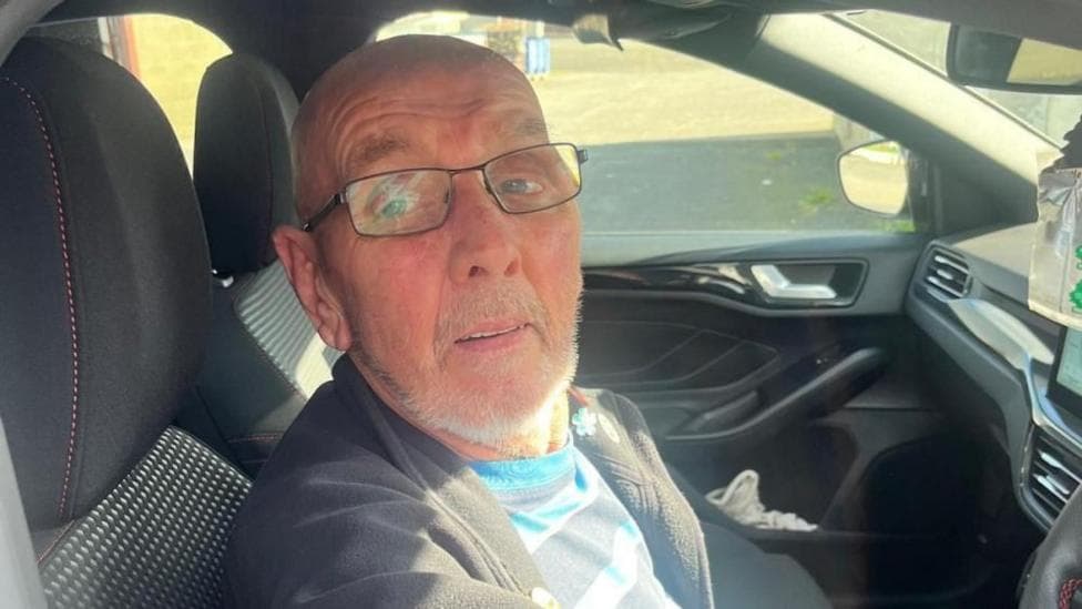 Reuben is bald and wears black glasses. He is wearing a blue striped t-shirt and grey jacket. He is sitting in a car.