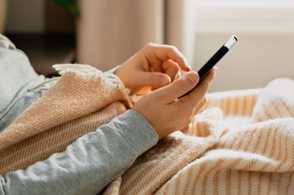 Close up of someone in bed holding a black phone, wearing a long grey jumper