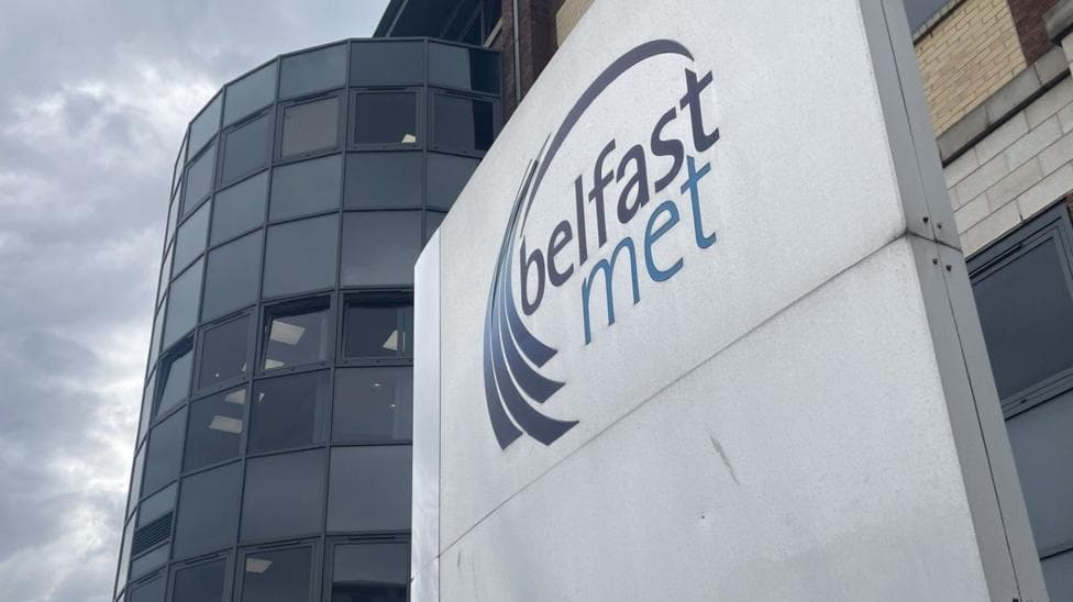 A beige brick building has a black window semi-cylinder entrance. It has a grey-metal sign in front of it that read "Belfast met" in blue writing.