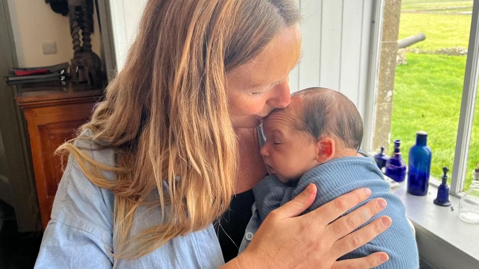 Mum Alice Traill Ford with baby Duncan Wilder Thorstein Traill Suarez Ford. She is wearing a blue overshirt and black top and kissing the top of the baby's head. The baby is wrapped in a blue blanket.