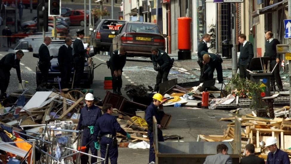 Security forces examine the scene of the Omagh bombing. There is rubble across the road and officers are wearing uniform and hard hats.