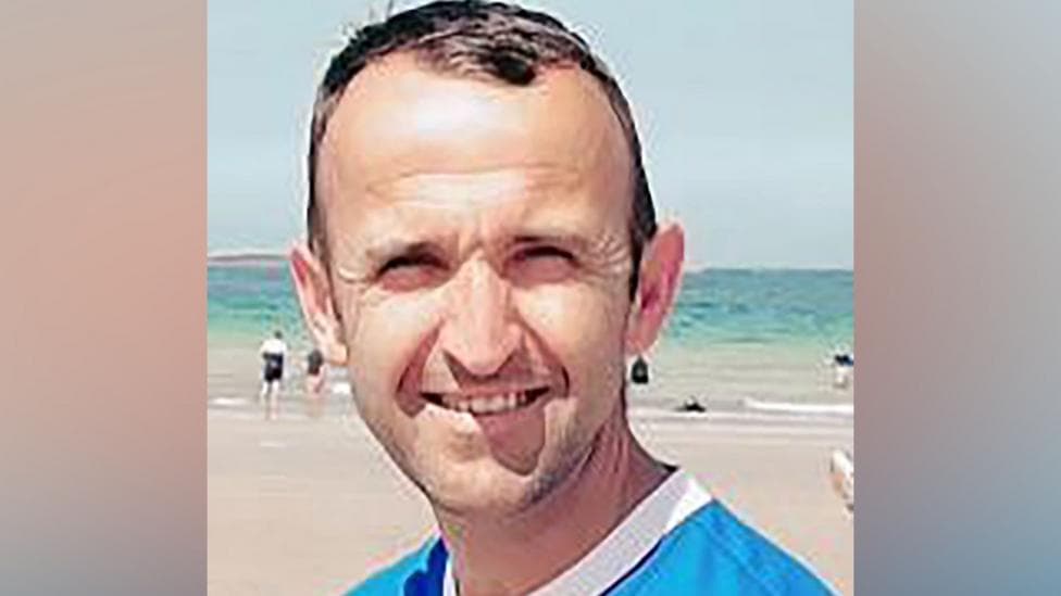 Christopher is smiling at the camera. He has short brown hair. He is wearing a blue top with a white collar.