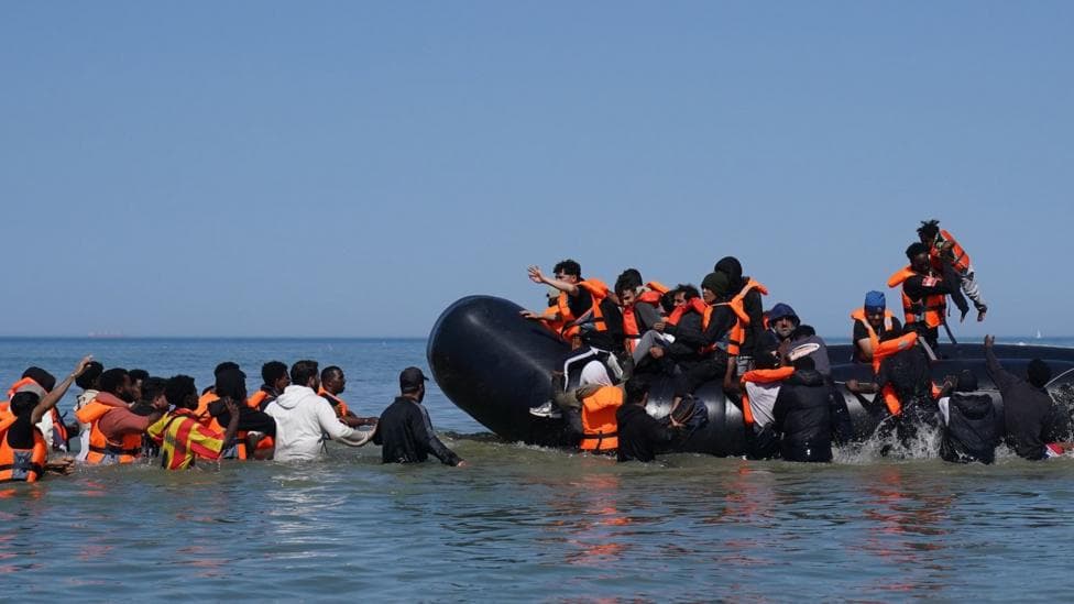 People thought to be migrants attempt to board a small boat off the coast of Hardelot, France, during an attempt to cross the English Channel on 29 July 2026. A group of people are on the inflatable vessel wearing orange life jackets and others are in the water, waiting to board the dinghy.