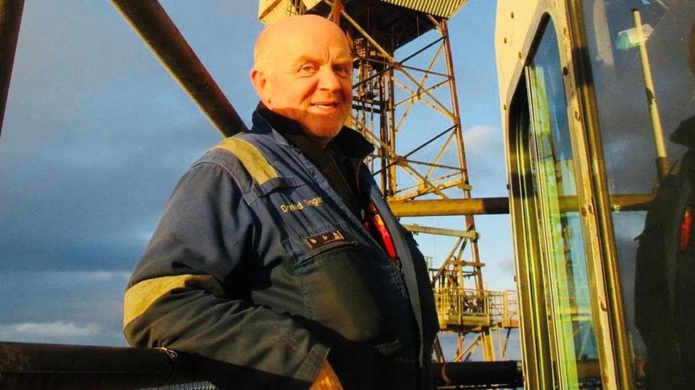 A man, Donald Singer, in blue overalls, smiling at the camera, on an offshore platform.