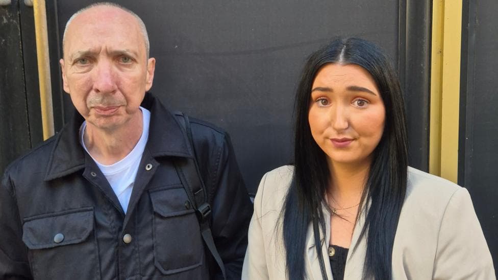 A man with grey hair and a black jacket, with a white shirt. He stands beside a woman with long black hair and a grey jacket.