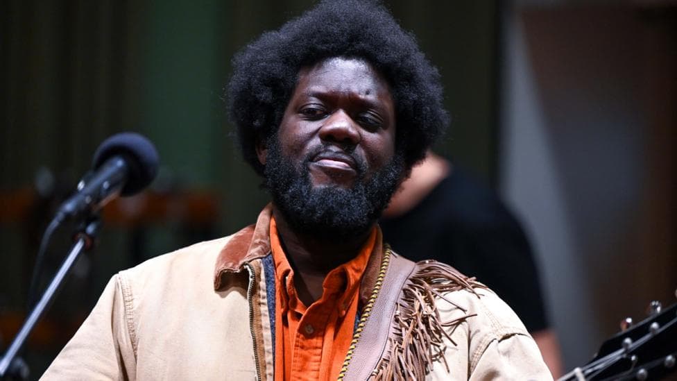 Michael Kiwanuka in a light brown jacket and orange shirt, holding a guitar and standing in front of a microphone