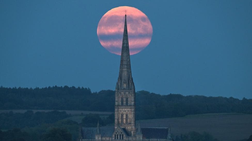 A pink-coloured full Harvest Moon rises behind Salisbury Cathedral spire. There are hills and trees in the distance and streaks of grey cloud across the Moon