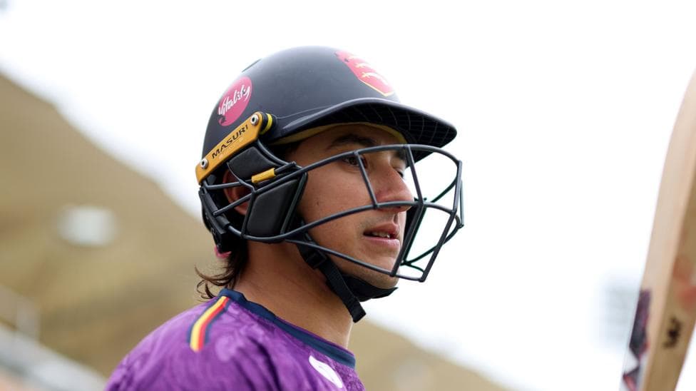 Robin Das wearing a black cricket helmet, carrying a cricket bat and wearing a purple top