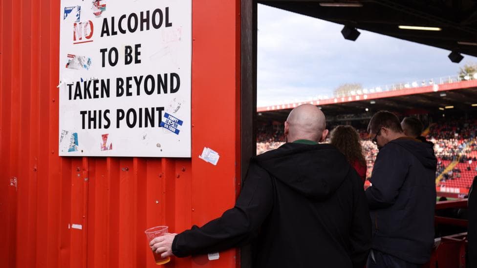 A football fan holds his pint of beer beneath a sign instructing that no alcohol should be taken beyond this point so he can continue to watch the match. The fan has his back to the camera and is wearing a dark-coloured jacket.