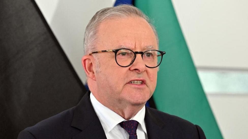 Australia's Prime Minister Anthony Albanese speaks at a news conference in front of several flags. He is wearing a dark suit with a red spotted tie and dark glasses