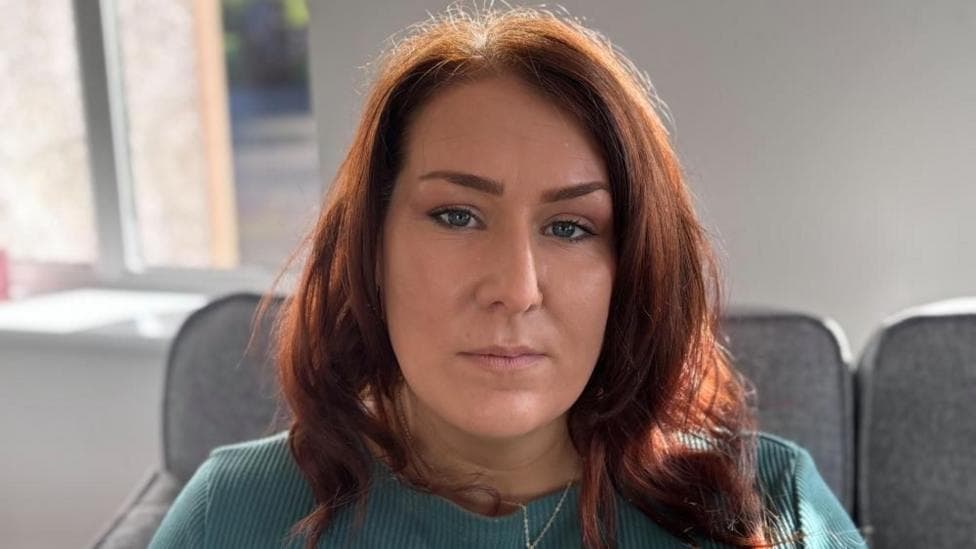 A woman with red hair looks into the camera. She is wearing a green top and is sitting on a grey sofa.